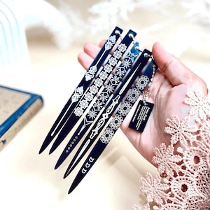 May include: A collection of six dark blue hair sticks with intricate white floral and geometric designs. Each stick is held in a hand, showcasing the variety of patterns. A small tag with text hangs from one stick.