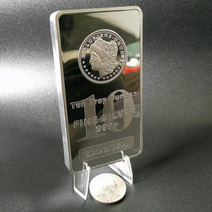 May include: A rectangular silver bar with a coin design on top, displaying the text "Ten Troy Ounces FINE SILVER 999+" and "Made in U.S.A." A silver coin rests below the bar. The bar is displayed on a clear stand.