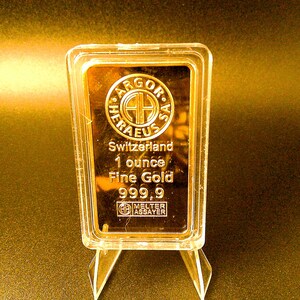 May include: A gold bar encased in clear plastic, displayed on a clear stand. The bar is stamped with the words "ARGOR-HERAEUS Switzerland 1 ounce Fine Gold 999.9" and a logo. The gold bar is approximately 1 inch in height.
