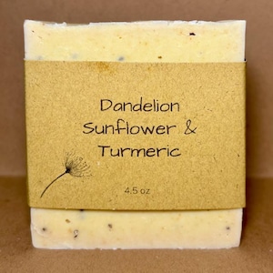 May include: A rectangular bar of soap with a light yellow hue, wrapped with a brown paper label. The label reads "Dandelion Sunflower & Turmeric" with a small illustration and "4.5 oz".
