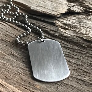 Könnte beinhalten: Ein silberfarbener Erkennungsmarken-Anhänger aus Metall mit gebürsteter Oberfläche, der an einer passenden Kugelkette hängt. Das rechteckige Schild hat abgerundete Ecken und eine glatte, metallische Oberflächenstruktur auf dem gesamten Stück.