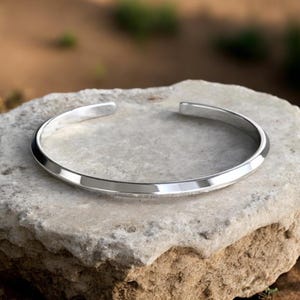 May include: A polished silver cuff bracelet. The bracelet has a smooth, rounded design with a slight curve. The open ends of the bracelet are visible. The bracelet is resting on a light grey stone.