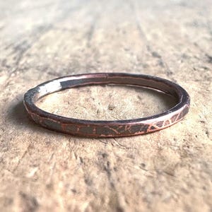 May include: A hammered copper ring with a textured surface. The ring has a dark, aged appearance, with variations in colour from reddish-brown to darker shades. The band is thin and appears handmade, showcasing a rustic, artisan style.