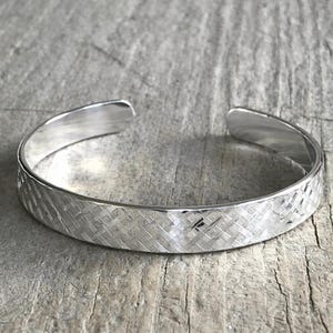 May include: A silver cuff bracelet with a textured, woven pattern. The open-ended bracelet is made of polished metal and sits on a wooden surface. The bracelet is approximately 2 cm wide.