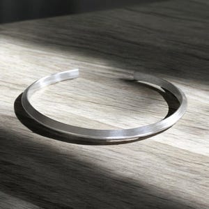 May include: A silver cuff bracelet with a simple, flat design. The open-ended bracelet is made of a smooth, metallic material. The jewellery is displayed on a wooden surface, with natural light creating shadows.