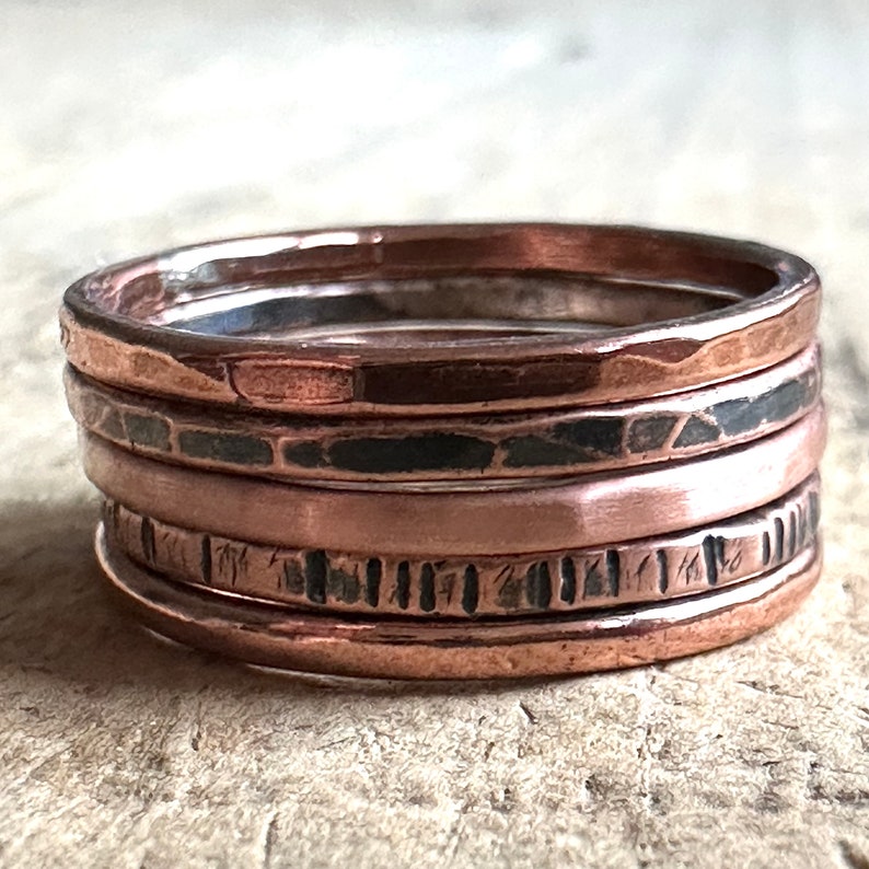 May include: Set of five copper rings, stacked on top of each other. The rings are hammered and have a rustic, worn look. The top ring is smooth, the second ring has a hammered texture, the third ring is smooth, the fourth ring has a hammered texture, and the bottom ring is smooth.