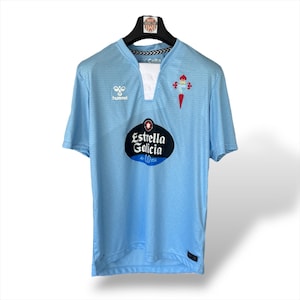 May include: Light blue soccer jersey with the text "Estrella Galicia" and the Hummel logo. The jersey has a V-neck design with a white insert and a team crest on the chest. The jersey is hanging on a black hanger.