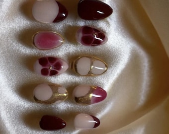 May include: Set of ten fake nails with various designs. The nails are oval shaped and feature a burgundy, white, and gold color scheme. Some nails have a floral design, while others have polka dots or a half-moon design.