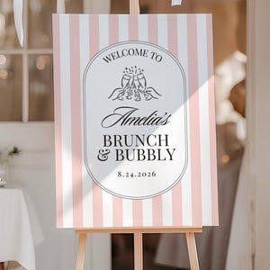 Puede incluir: Un caballete de madera exhibe un letrero con rayas rosas y blancas. El letrero dice "Welcome to Amelia's Brunch & Bubbly 8.24.2026" en una escritura elegante y un emblema decorativo. Globos blancos y cintas adornan el fondo.