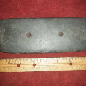 May include: A gray stone tool with two holes drilled into it. The tool is approximately 6 inches long and is resting on a red surface. A wooden ruler is below the tool for scale.