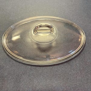 Puede incluir: Una tapa de vidrio transparente de forma ovalada con un asa de metal. La tapa está diseñada para utensilios de cocina y presenta un asa elevada para facilitar el levantamiento. El diseño transparente permite la visibilidad del contenido.