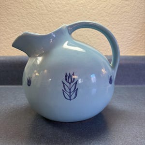Può includere: Una brocca in ceramica azzurra con un corpo arrotondato e un manico curvo. La brocca è dotata di un beccuccio per versare ed è decorata con disegni di tulipani blu scuro. La brocca è un oggetto decorativo per servire bevande.