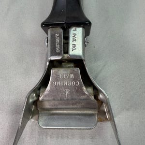 Może przedstawiać: Zbliżenie uchwytu do garnka Corning Ware. Uchwyt jest czarny i przymocowany do srebrnej metalowej ramy. Rama ma nadrukowany tekst "CORNING WARE" oraz "PAT. NO." i "2,851,293".