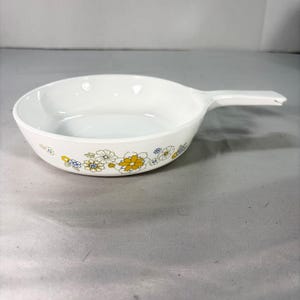 May include: White ceramic skillet with a single handle and a floral pattern. The floral design features yellow, blue, and white flowers. The skillet is designed for cooking and serving.
