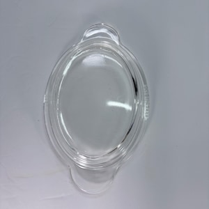 Puede incluir: Tapa ovalada transparente de Pyrex con dos asas pequeñas. La tapa es de vidrio transparente y tiene un borde elevado. El logotipo de Pyrex es visible en el lateral. Esta tapa está diseñada para utensilios de cocina.