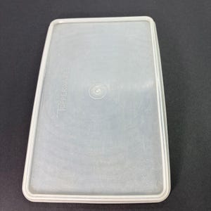 May include: A clear, rectangular Tupperware lid with a slightly textured surface. The lid has a raised edge and the Tupperware brand name is visible. The lid is designed to fit a rectangular container.
