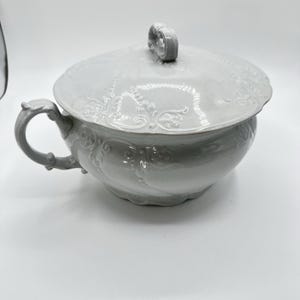 Op de afbeelding: Een witte keramische soepterrine met deksel en handvat. De terrine heeft een ronde body met sierlijke bloemen- en krulwerkdetails. Het deksel heeft een decoratieve handgreep en een bijpassend reliëf. De handgreep heeft een gebogen, lusvorm.