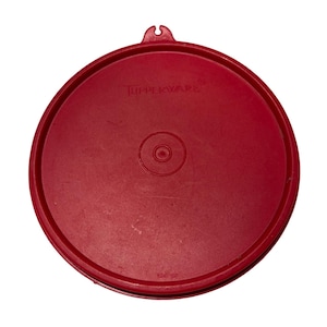 以下が含まれることがあります： 中央に円形のデザインと小さなハンドルが付いた、丸い赤いTupperwareの蓋。「Tupperware」の文字が蓋に印刷されています。蓋は耐久性のあるプラスチック素材で作られています。