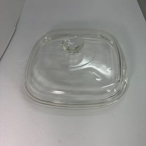 May include: A clear, square glass lid with a small, round handle in the center. The lid has rounded corners and a slightly raised edge. The glass is transparent, allowing a view of what it covers. This is a kitchenware item.