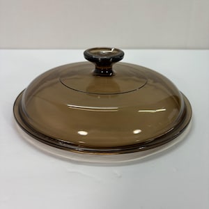 Puede incluir: Una tapa de vidrio transparente marrón con forma redondeada y un pequeño pomo marrón oscuro en la parte superior. La tapa está diseñada para encajar en una olla o sartén y tiene un borde ligeramente elevado.