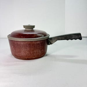 Op de afbeelding: Een vintage kastanjebruine steelpan met een deksel en een lange, zwarte handgreep. De pot heeft een zilveren rand en een bijpassend deksel met een kleine, ronde knop. De handgreep is getextureerd voor grip. Dit kookgerei is geschikt om te koken.