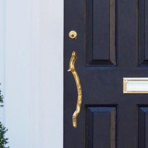 May include: A black front door with gold hardware, including a door knocker, handle, and mail slot. The door has six rectangular panels and is set against a white wall. The text "Nakhi MA" is visible at the bottom of the door.