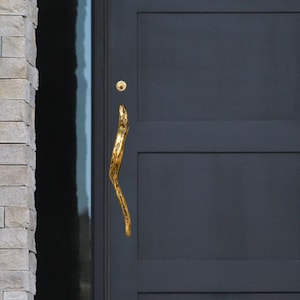 Könnte beinhalten: Eine dunkelgraue Eingangstür mit einem goldfarbenen, organisch geformten Griff. Die Tür hat ein rechteckiges Paneeldesign. Hellfarbene Steinmauern und schwarze Pflanzgefäße mit Grün umrahmen die Tür. Der Text "Nakhi MA" ist unten sichtbar.