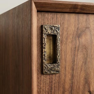 Può includere: Primo piano di un mobile in legno con una maniglia incassata rettangolare color ottone. La maniglia presenta un design ornato e strutturato. Il mobile è in legno marrone scuro con venature visibili.