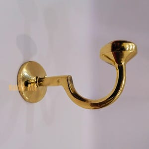 Könnte beinhalten: Ein goldfarbener Wandhaken mit kreisförmiger Basis und gebogenem Haken. Der Haken hat ein abgerundetes Ende und ist auf einer weißen Oberfläche montiert. Der Haken ist aus Metall und hat eine glänzende Oberfläche.