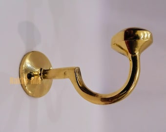 May include: A gold-colored wall hook with a circular base and a curved hook design. The hook has a rounded end and is mounted on a white surface. The hook is made of metal and has a shiny finish.