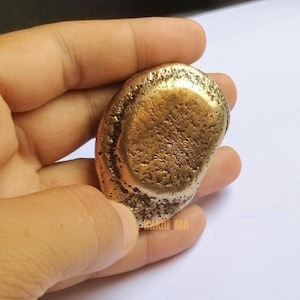 May include: A golden-colored, irregularly shaped object with a textured surface. The object is held in a hand against a white background. The object appears to be made of metal.