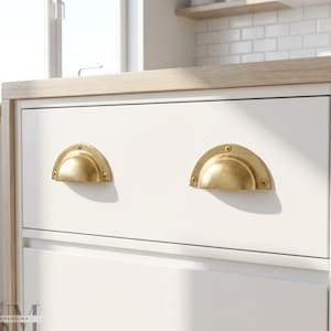 May include: Close-up of a white cabinet with two brass-colored, half-moon drawer pulls. The cabinet has a light wood countertop and is in a kitchen setting. The drawer pulls are mounted with small screws.
