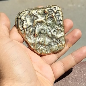 May include: A gold-colored, irregularly shaped object with a textured surface. The object is held in a hand, and appears to be a decorative piece or a small sculpture. The surface has a rough, uneven appearance.