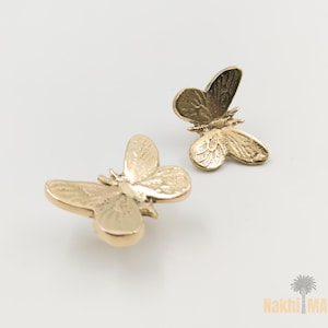 May include: Two gold-colored butterfly-shaped decorative objects. The butterflies have detailed wing patterns and are positioned on a white background. The objects appear to be made of metal and are likely used for decorative purposes.