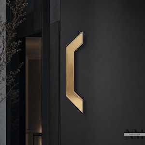 Può includere: Una maniglia per porta geometrica e angolare di colore dorato montata su una porta nera opaca. La maniglia presenta una forma distintiva a chevron con angoli acuti e una finitura metallica liscia. Il testo NAKHILMA è visibile nell'angolo in basso a destra.