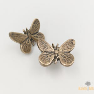 May include: Two bronze-colored butterfly-shaped decorative objects. The butterflies have detailed wing patterns and are positioned on a white background. The objects appear to be made of metal and are likely used for decorative purposes.