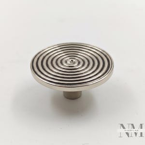 Puede incluir: Un tirador de armario plateado con un diseño en espiral. La cara circular tiene ranuras concéntricas y un vástago cilíndrico. El diseño recuerda a un objetivo. El tirador es probablemente de metal y adecuado para muebles. Diámetro aproximado de 4 cm.