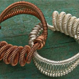Op de afbeelding: Twee handgemaakte draadarmbanden. Eén is koperkleurig en de andere zilverkleurig. De armbanden zijn met elkaar verbonden. De koperen armband bevindt zich links en de zilveren armband rechts. De armbanden liggen op een blauwgroen houten oppervlak.