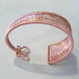 Op de afbeelding: Een koperkleurige draadmanchetarmband. De armband heeft een geweven ontwerp met verticale lijnen en een haaksluiting. Het open manchetontwerp maakt het gemakkelijk om te dragen. De armband is een uniek sieraad.