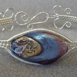 以下が含まれることがあります： 銀色のワイヤーラップブレスレット。ダイクロイックガラスの中央部分があり、青、紫、金色の配色です。ブレスレットはフッククラスプと装飾的な渦巻きが特徴です。