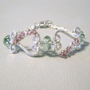 May include: A silver wire bracelet with heart-shaped elements. It features light green and pink gemstones, with a central green stone. The bracelet has a delicate, handcrafted appearance, suitable for everyday wear or special occasions.
