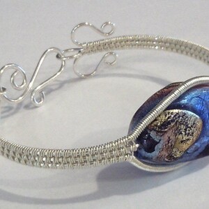 以下が含まれることがあります： 銀色のワイヤーブレスレット。ダイクロガラスビーズが特徴で、青、紫、金の混合色です。ブレスレットは編み込まれたワイヤーデザインと装飾的な留め金が付いています。ユニークなジュエリーです。
