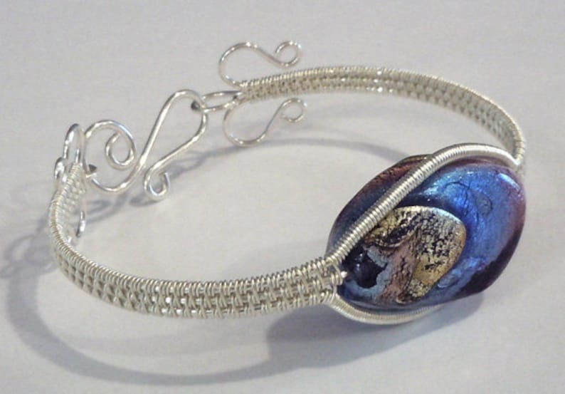 以下が含まれることがあります： 銀色のワイヤーブレスレット。ダイクロガラスビーズが特徴で、青、紫、金の混合色です。ブレスレットは編み込まれたワイヤーデザインと装飾的な留め金が付いています。ユニークなジュエリーです。