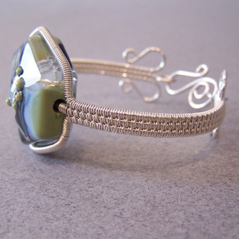 以下が含まれることがあります： 大きなファセットガラスビーズが付いた、銀色のワイヤーラップブレスレット。ビーズは緑と青の色合いが特徴です。ブレスレットは織り込まれたワイヤーバンドと、端に装飾的な渦巻き模様があります。