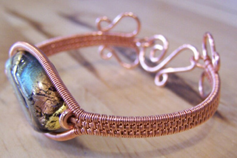 以下が含まれることがあります： 織りバンドと装飾的な渦巻き模様の銅線ブレスレット。ブレスレットは、金と青の色合いの長方形の二色性ガラスビーズが特徴です。金属細工は複雑で詳細で、職人のスキルを示しています。