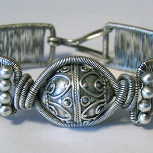 Puede incluir: Pulsera plateada con un elemento decorativo central. La pulsera presenta un dise&ntilde;o de alambre tejido en la banda y una pieza central detallada y estampada. Peque&ntilde;as cuentas plateadas acent&uacute;an los lados y el cierre es visible.