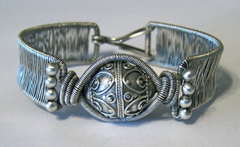 Puede incluir: Pulsera plateada con un elemento decorativo central. La pulsera presenta un dise&ntilde;o de alambre tejido en la banda y una pieza central detallada y estampada. Peque&ntilde;as cuentas plateadas acent&uacute;an los lados y el cierre es visible.