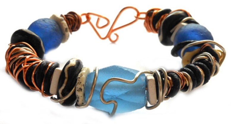 Pode incluir: Pulseira artesanal com contas azuis, pretas e brancas, com detalhes em fio de cobre e prata. A pulseira inclui uma grande conta facetada azul clara no centro. Fecho de gancho e argola em cobre.