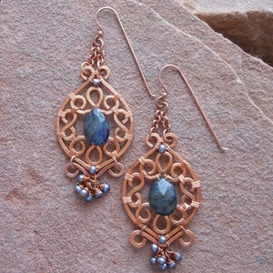 May include: Copper wire earrings with a filigree design, featuring a teardrop-shaped blue-green stone in the center and small blue beads at the bottom. The earrings have a hook closure.