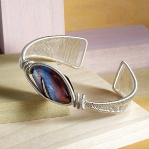 Puede incluir: Una pulsera de pu&ntilde;o de alambre plateado con una cuenta central de forma ovalada. La cuenta presenta una mezcla de colores azul y granate. El dise&ntilde;o de la pulsera incorpora un intrincado enrollado de alambre y detalles calados.